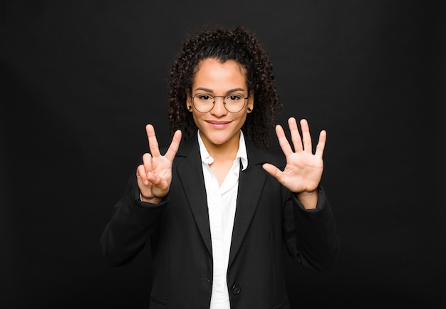 Young black woman smiling and looking friendly, showing number seven or seventh with hand forward, counting down against black wall