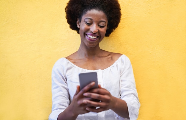 Giovane donna di colore che sta isolata sopra giallo facendo uso del telefono cellulare astuto