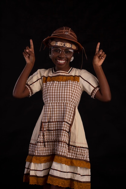 Young black woman wearing glasses and party dress with\
cancageiro hat and studio photo with black background festa\
junina