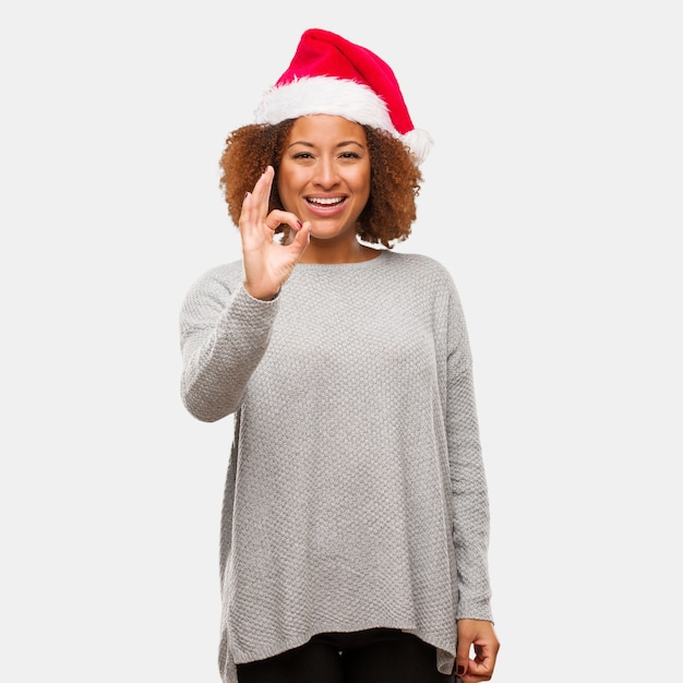 若い黒の女性は、サンタの帽子を着て明るく、自信を持ってOKジェスチャー