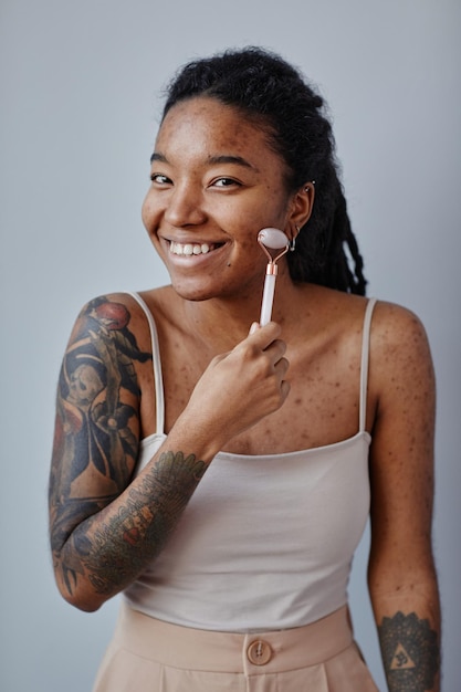 Young black woman with acne scars and tattoos using face massager and smiling