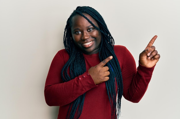 Young black woman with braids wearing casual clothes smiling and looking at the camera pointing with two hands and fingers to the side