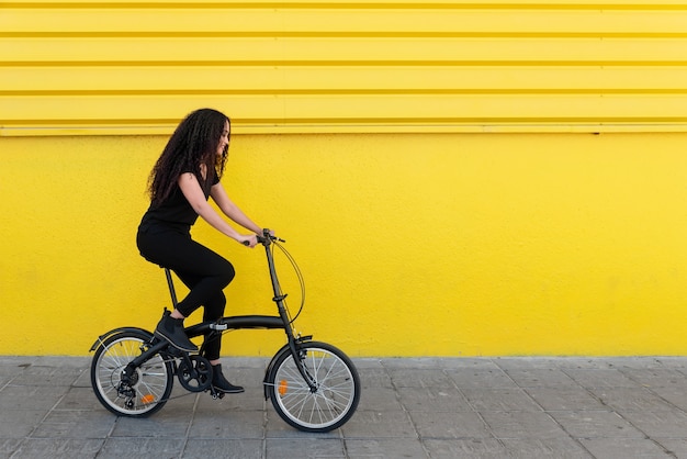 黄色い壁の前で彼女の自転車に乗って長い髪と黒い服を着た若い黒人女性