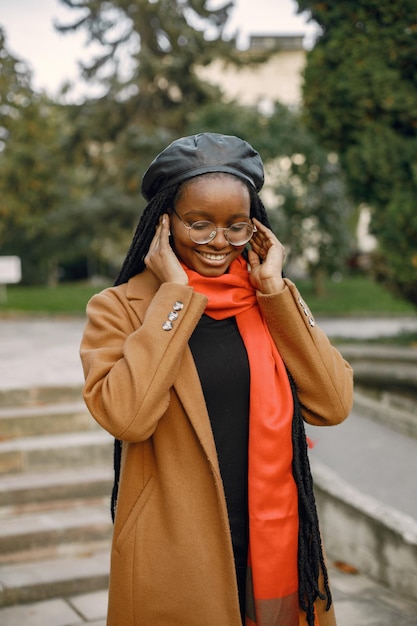 Young black woman with a long locs hairstyles standing outside. Woman wearing brown coat, orange scarf and black hat