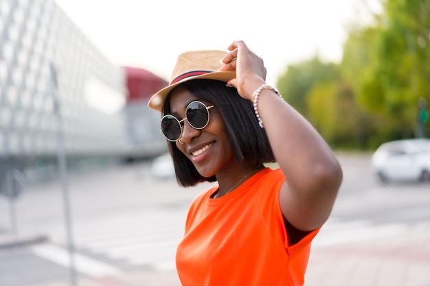 Young black woman with sunglasses and hat wearing orange tshirt smiling in the city during summer lifestyle photos