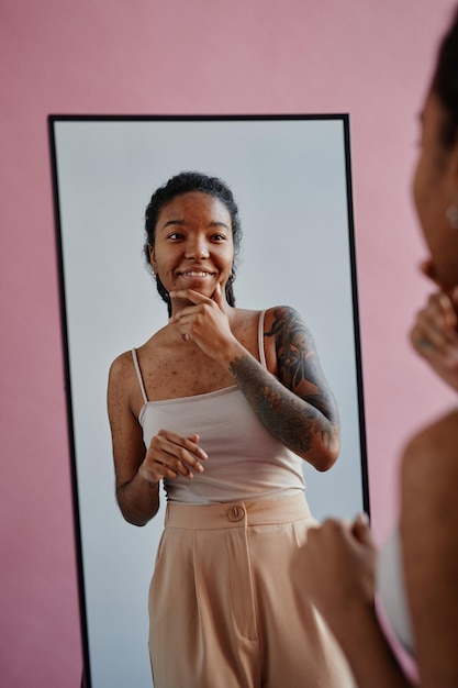 Young black woman with tattoos and acne scars looking in mirror