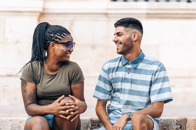 Young black woman with tattoos and sunglasses sitting next to a caucasian man laughing in the street