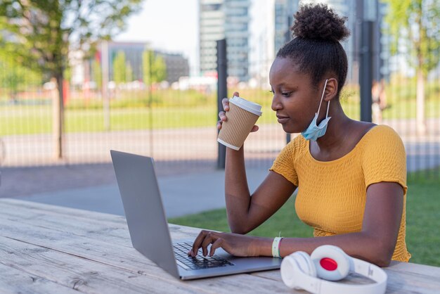 カプチーノコーヒーをすすりながら公園で保護マスクの若い学生の屋外での流行の時に彼女のラップトップで仕事をしている若い黒人女性