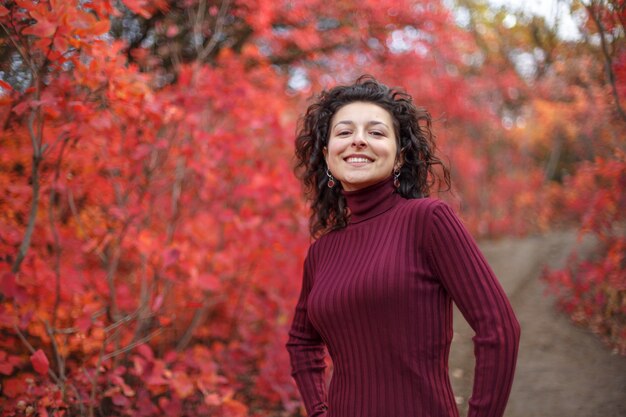 Young blackhaired woman in red sweather posing on camera in red autumnn bushes.