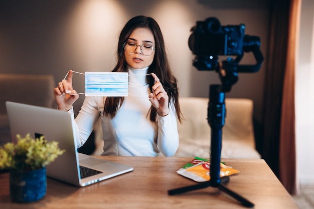 Young blogger in glasses female is filming her vlog and showing her audience to be safe and use medical mask. Coronovirus safe workplace concept. Putting medical mask on face.