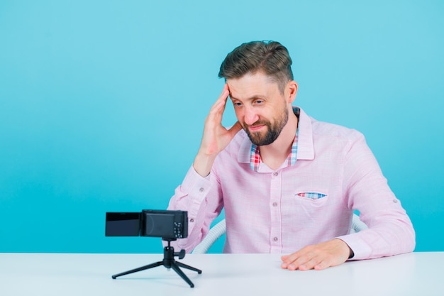 Young blogger man is holding hand on temple by sitting in front of his mini camera on blue background