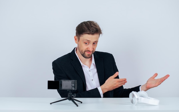 Young blogger man is showing right with hand by sitting in front of his mini camera on white background