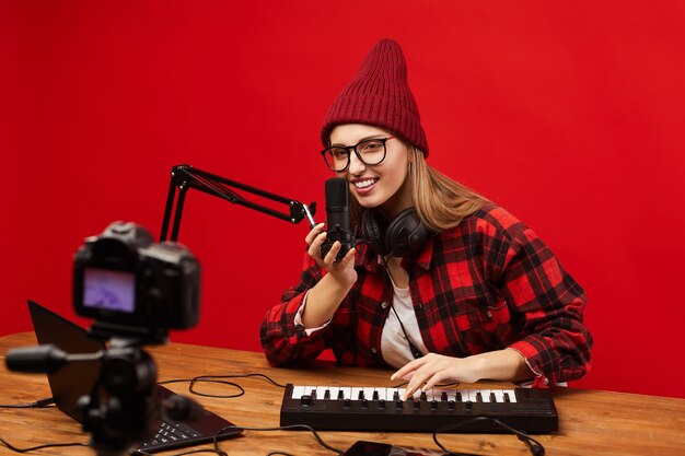 Giovane blogger seduta al tavolo che suona sulla tastiera musicale e canta nel microfono che gira video per i suoi follower