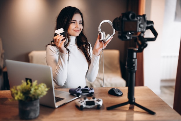 Молодая женщина-блогер с ноутбуком и джойстиками снимает видео и демонстрирует свое предпочтение в наушниках для видеоигр. Влиятельная молодая женщина в прямом эфире дома.