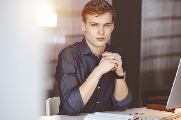 Фото Молодой блондин-бизнесмен думает о стратегии на своем рабочем месте с компьютером в затемненном офисе, яркий свет на заднем плане