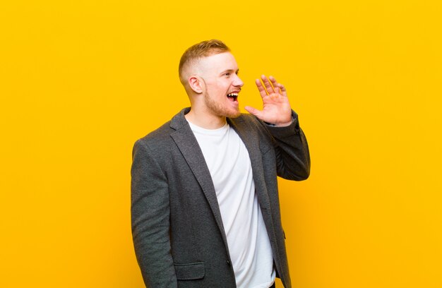 Young blond businessman yelling loudly and angrily to copy space on the side, with hand next to mouth