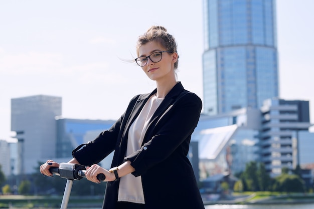 Young blond businesswoman in formalwear and eyeglasses standing on electric scooter against modern architecture on sunny day