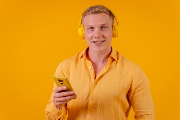 Young blond caucasian man on a yellow background with a cell phone and wireless headphones listening to music