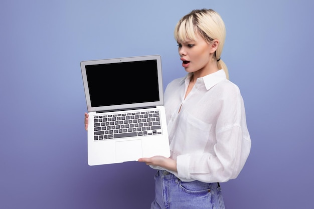 Young blond caucasian woman with a short haircut dressed in a white shirt and jeans holds a laptop