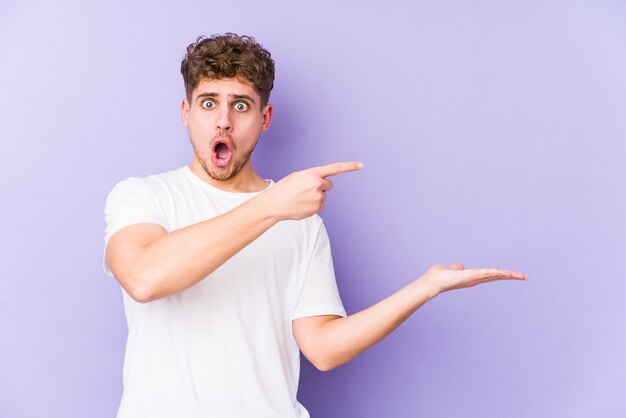 Young blond curly hair caucasian man excited holding a blank space on palm.