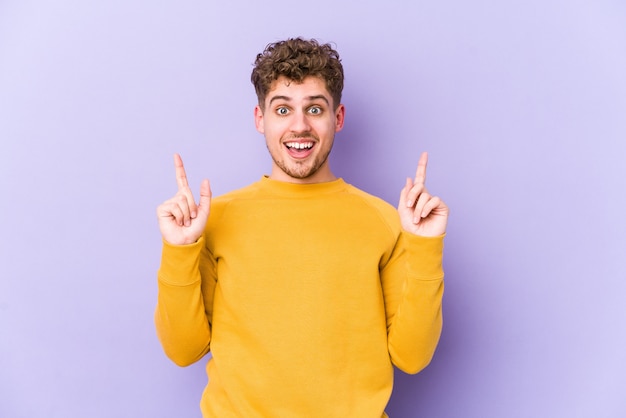 Young blond curly hair caucasian man isolated indicates with both fore fingers up showing a blank space.