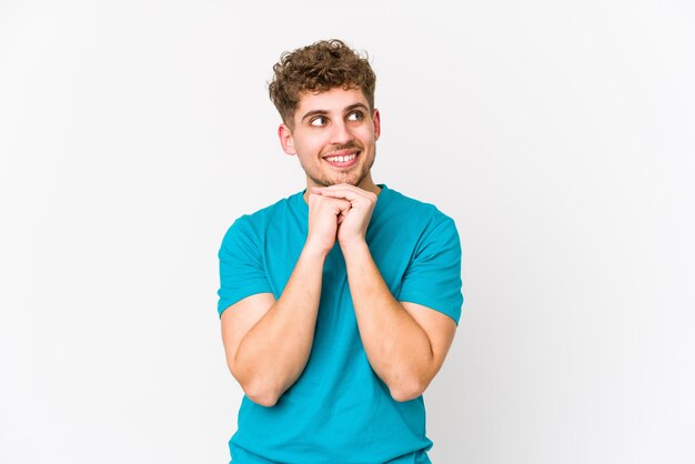 L'uomo caucasico dei giovani capelli ricci biondi isolato tiene le mani sotto il mento, sta guardando felicemente da parte.