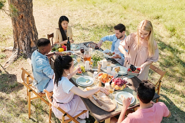 Young blond female in casual dress taking some cooked vegetables from bowl while standing by served table during dinner with her friends