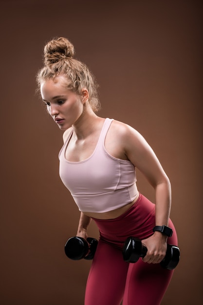 Young blond female in tracksuit bending slightly forwards while doing exercise for arms with dumbbells during workout in isolation