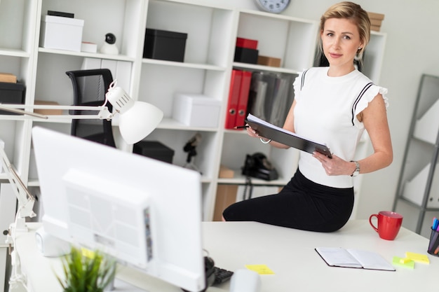 A young blond girl is sitting at the desk in the office and is holding documents and a pencil.