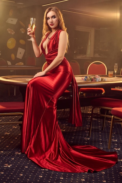 Young blond girl in a long red satin dress, with a glass of
champagne in her hand is posing sideways sitting on a poker table
in luxury casino. passion, cards, chips, alcohol, win, gambling -
it is a