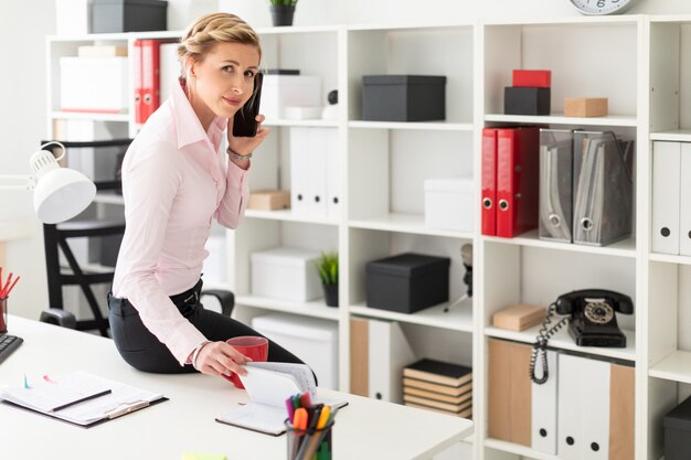 Una giovane ragazza bionda si siede sulla scrivania in ufficio, parla al telefono e sfoglia il blocco note.