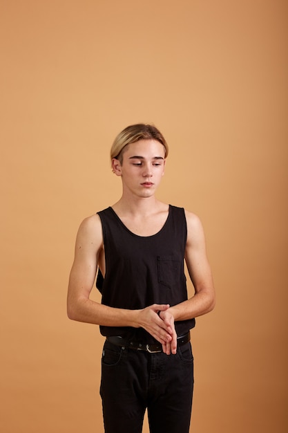 Молодой блондин парень, одетый в черную футболку и брюки, позирует в студии на бежевом фоне.