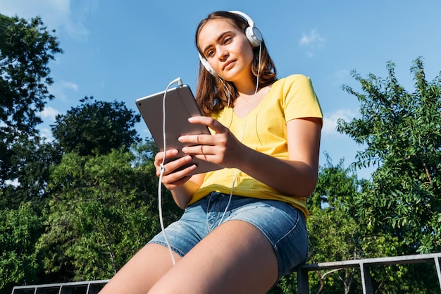 Young blond hair girl wearing yellow t-shirt and white headphones, smiling and looking at her tablet, sitting on a railing. Technology, lifestyle and relaxation concept