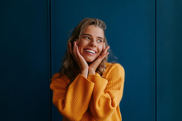 Фото Молодая блондинка в желтом свитере прикасается к лицу и улыбается, стоя на синем фоне