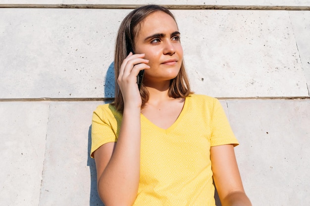 Young blond-haired girl wearing yellow t-shirt, talking with her smart phone leaning against a stone wall. Technology, music, lifestyle and relaxation concept.