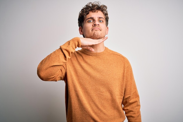 Young blond handsome man with curly hair wearing casual sweater over white background cutting throat with hand as knife threaten aggression with furious violence