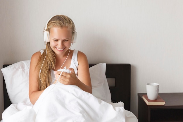 젊은 금발과 행복한 웃는 여성은 아침 시간에 흰색 침대에서 음악을 듣고 커피를 마십니다.