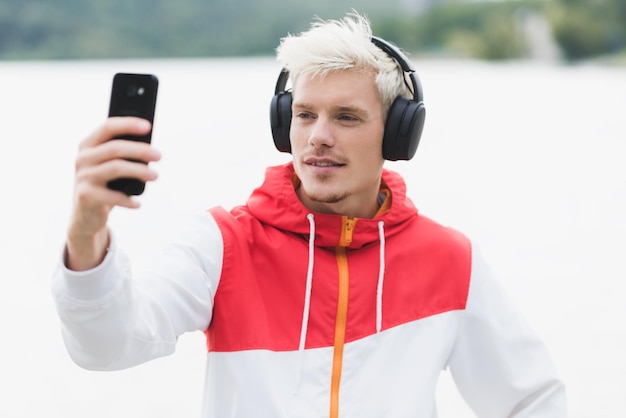최신 유행하는 빨간 지퍼 후드티를 입고 블루투스 헤드폰으로 음악을 듣는 젊은 금발 힙스터 남자는 야외에서 스마트 폰으로 셀카를 찍는 재미를 가지고 있습니다. 사람들의 라이프 스타일 기술 개념