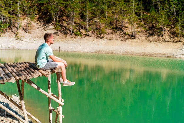 澄んだ水と緑の森の景色と山の湖の上の橋の上の若い金髪の男