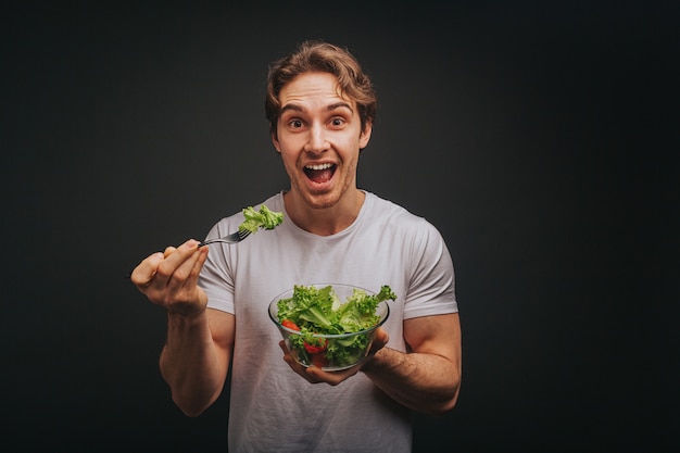 Young blond man in white t-shirt is holding happily plate of salad