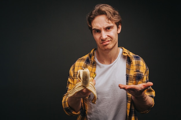 Young blond man in yellow shirt is doubtfully eating banana