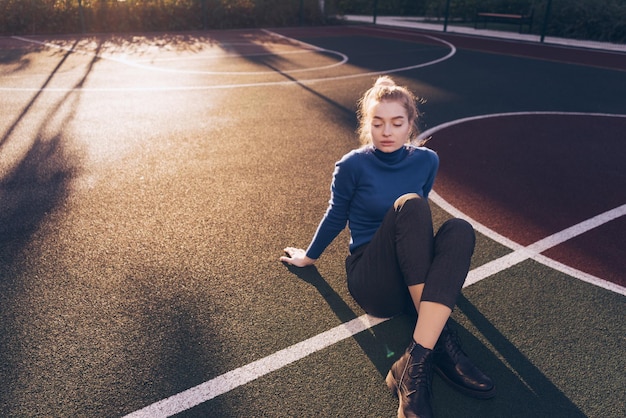 Young blond model girl in blue trendy sweater sits on the ground at a sports field posing