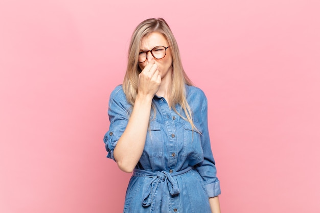 Young blond pretty woman feeling disgusted, holding nose to avoid smelling a foul and unpleasant stench