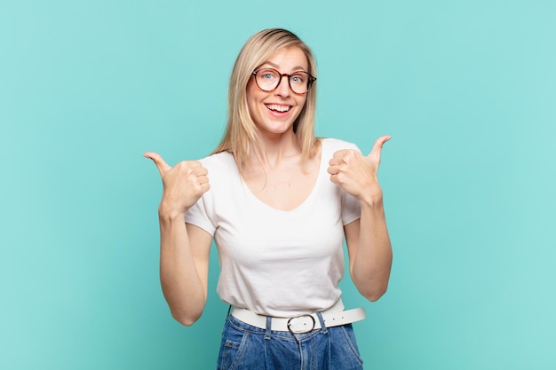 Young blond pretty woman smiling joyfully and looking happy, feeling carefree and positive with both thumbs up