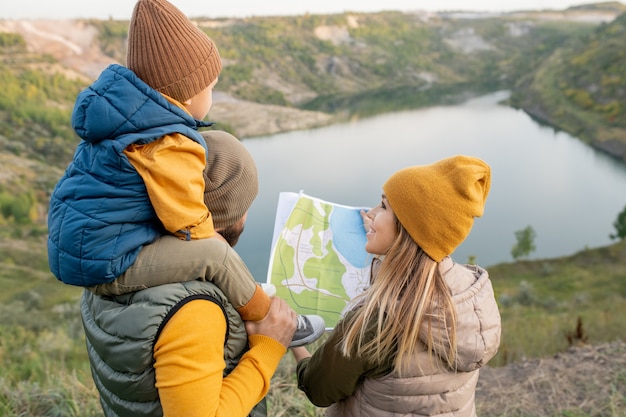 Young blond smiling woman with map looking at her husband with little son on neck standing in front of lake and deciding where to go