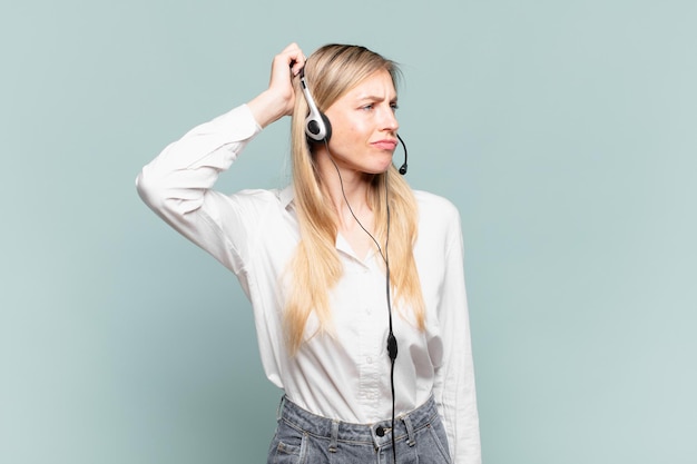 Young blond telemarketer woman feeling puzzled and confused, scratching head and looking to the side