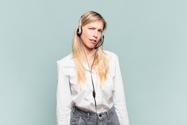 Young blond telemarketer woman feeling puzzled and confused, with a dumb, stunned expression looking at something unexpected