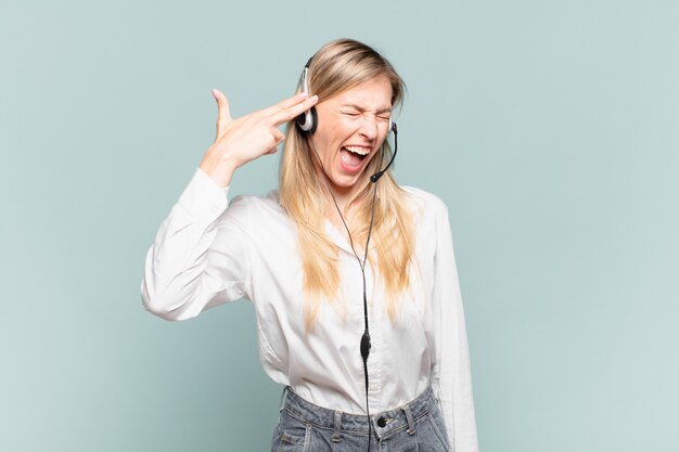 Young blond telemarketer woman looking unhappy and stressed, suicide gesture making gun sign with hand, pointing to head