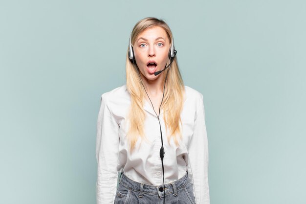 Young blond telemarketer woman looking very shocked or surprised, staring with open mouth saying wow