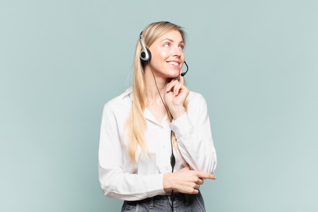 Young blond telemarketer woman smiling happily and daydreaming or doubting, looking to the side